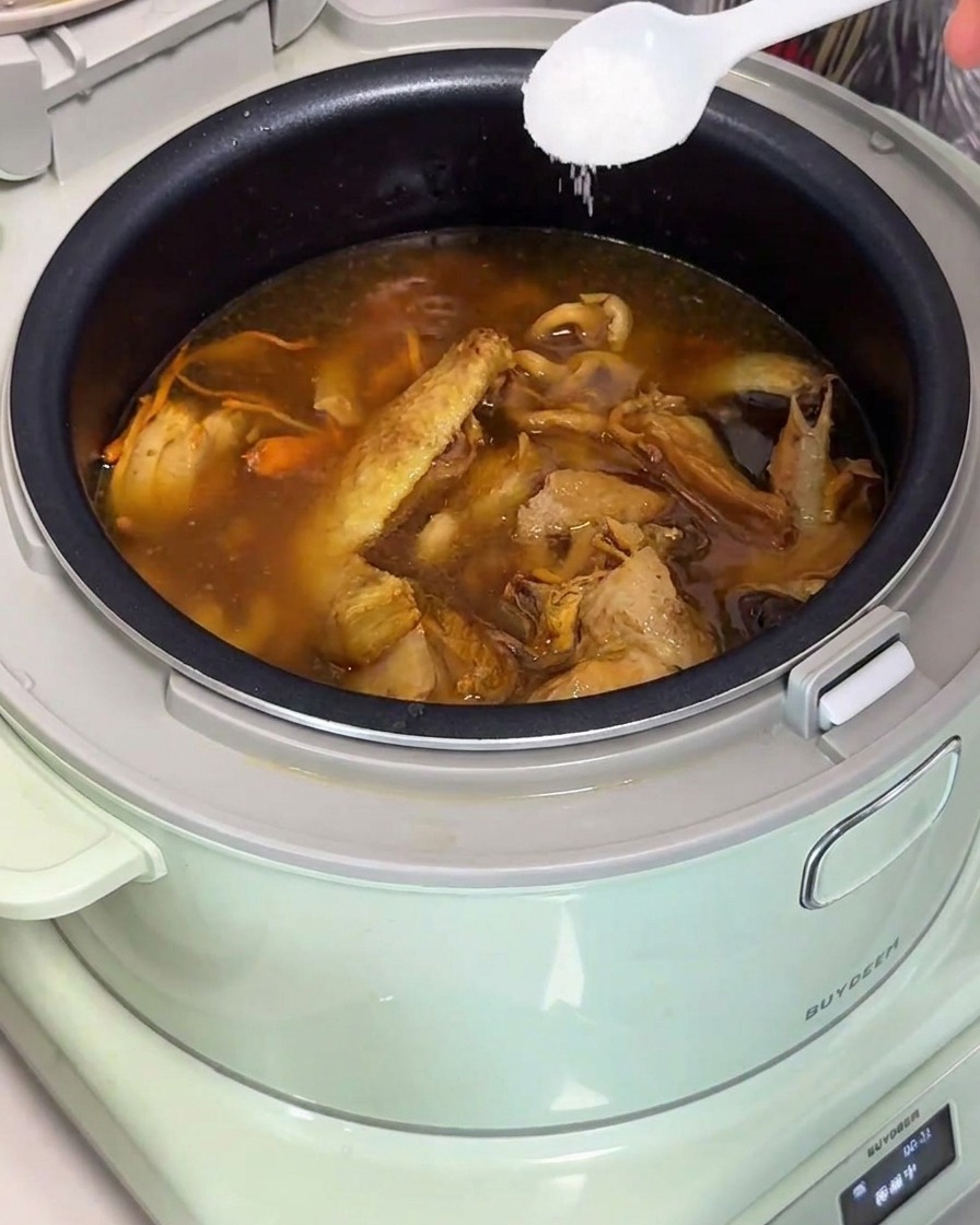 밥솥에 완성된 황금빛 닭고기 버섯 수프에 하얀 숟가락으로 소금을 넣는 모습.