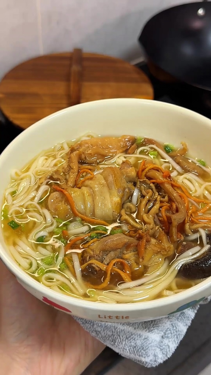 다양한 건버섯과 신선한 파가 곁들여진 김이 모락모락 나는 황금빛 닭고기 버섯 국수 한 그릇입니다.