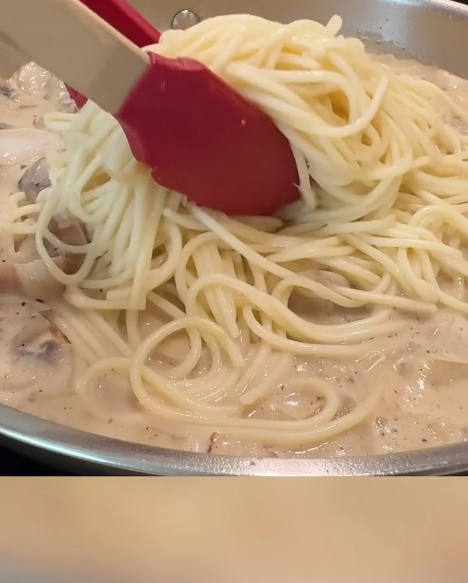 집게를 사용하여 파스타 면과 소스, 베이컨, 버섯을 고르게 섞고 있는 모습.