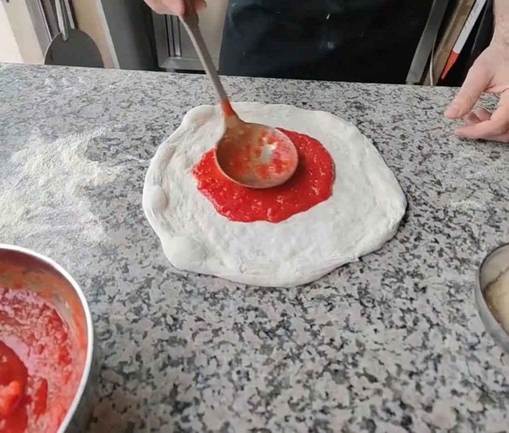 국자로 도우 중앙에 붉은 토마토 소스를 얹어 원을 그리며 펴 바르는 모습.