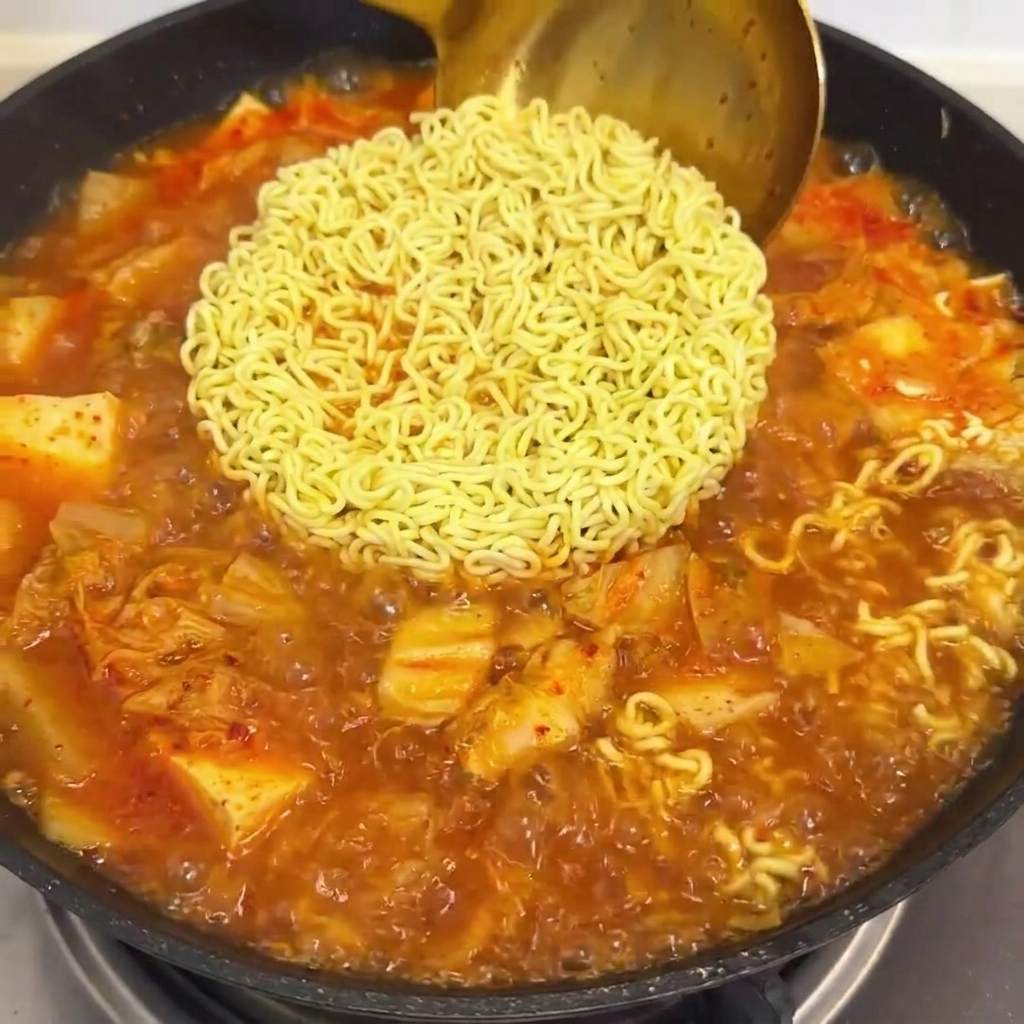 라면과 김치 국물이 끓고 있는 팬에 붉은 스프를 붓는 모습.