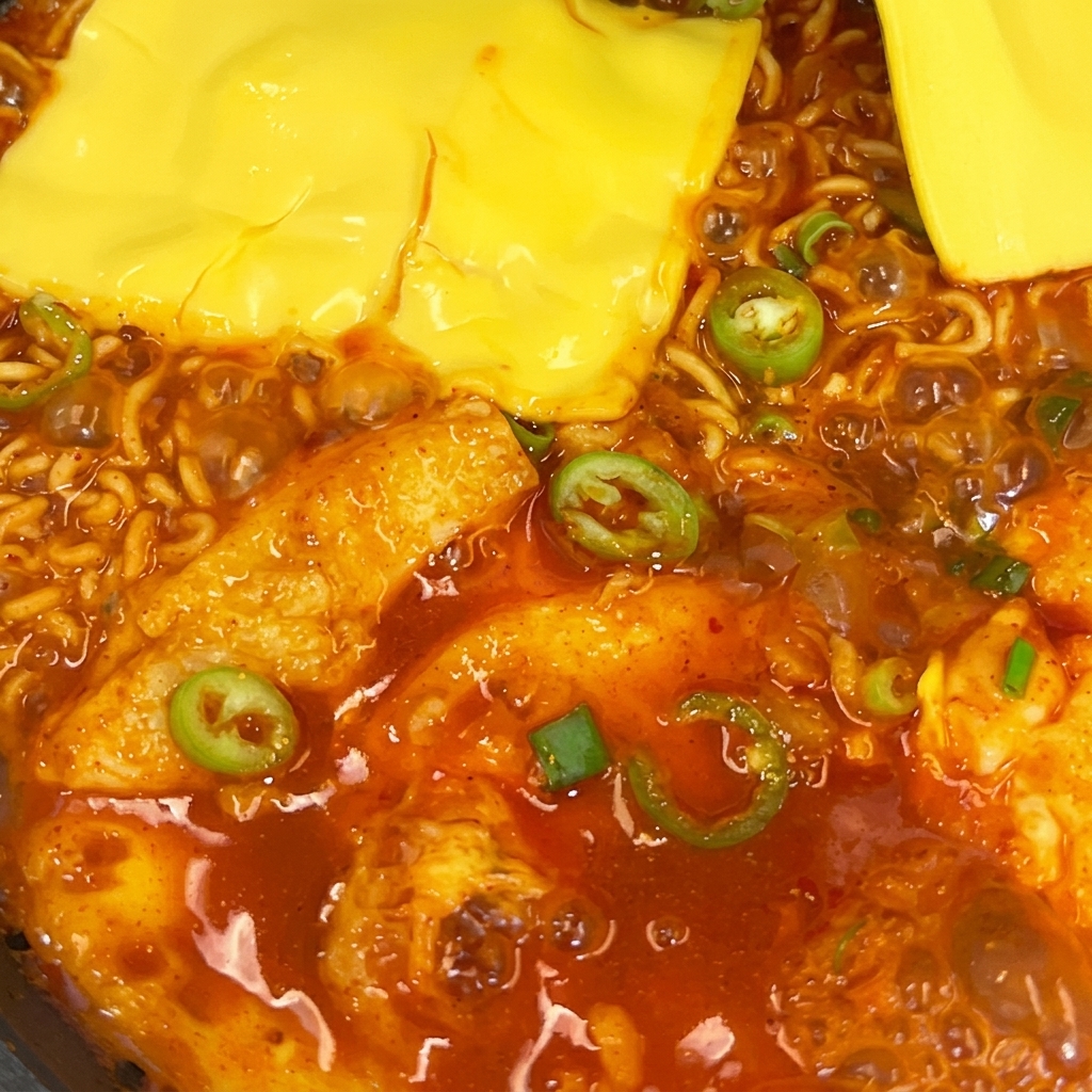 팬에서 끓고 있는 매운 라면 위에 슬라이스 치즈 두 장이 녹아내리는 모습.