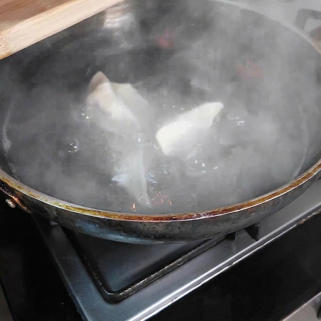 커다란 냄비 속 끓는 물 안에서 수증기가 모락모락 피어오르는 가운데 여러 개의 만두가 잠겨 있는 모습.