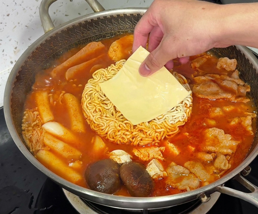 김이 모락모락 나는 부대찌개 안에서 라면 면발 위로 노란 치즈 슬라이스가 녹아내리는 모습.