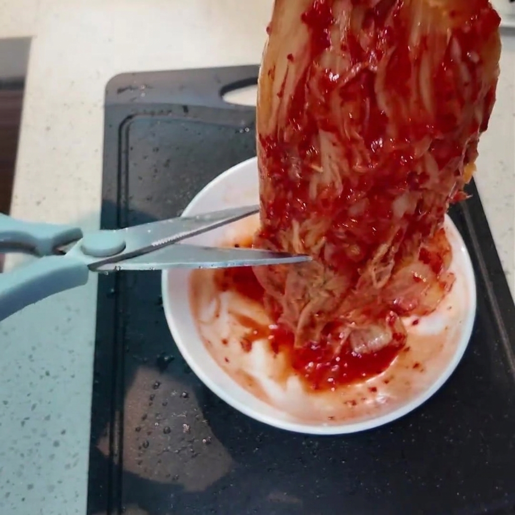 작은 흰 그릇 위에서 주방 가위로 빨간 김치를 작게 자르는 모습.