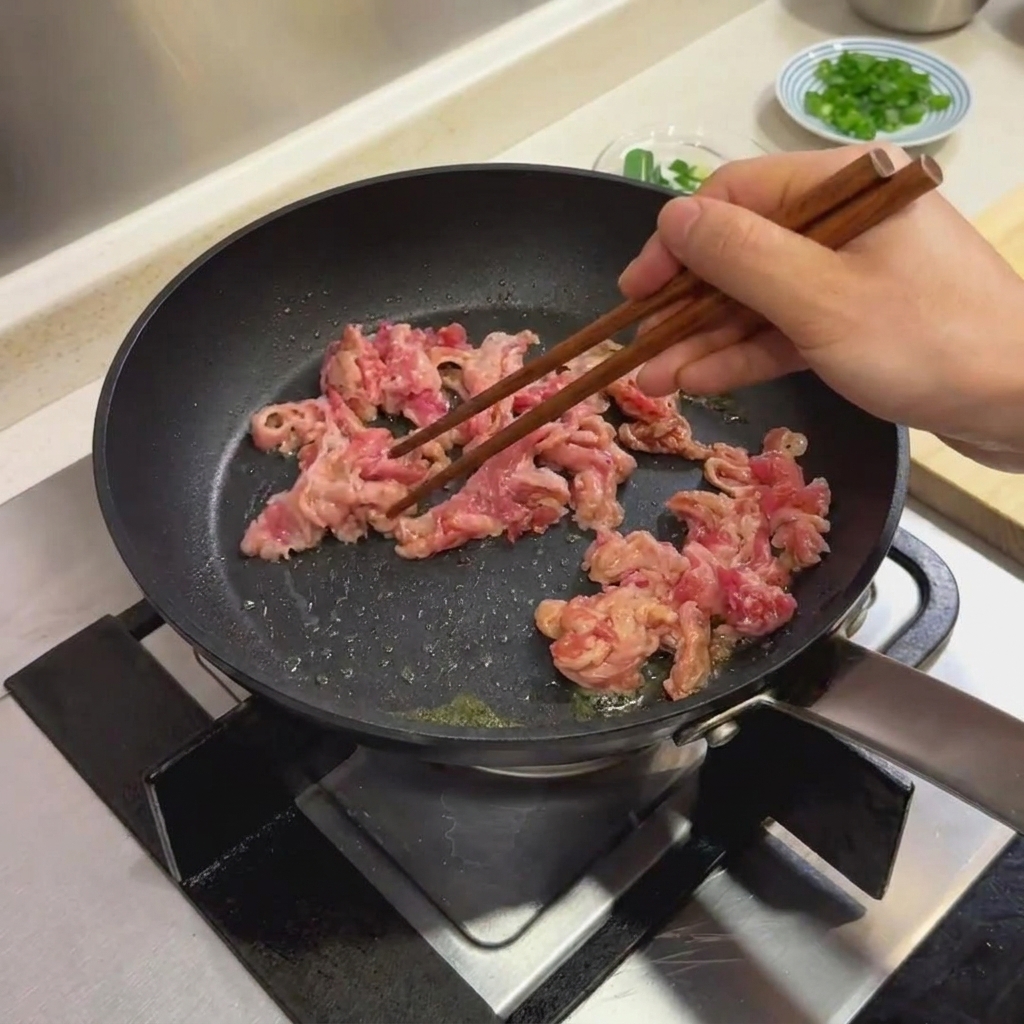 검은색 프라이팬에서 나무 젓가락으로 마리네이드한 소고기 슬라이스를 조리하는 모습.
