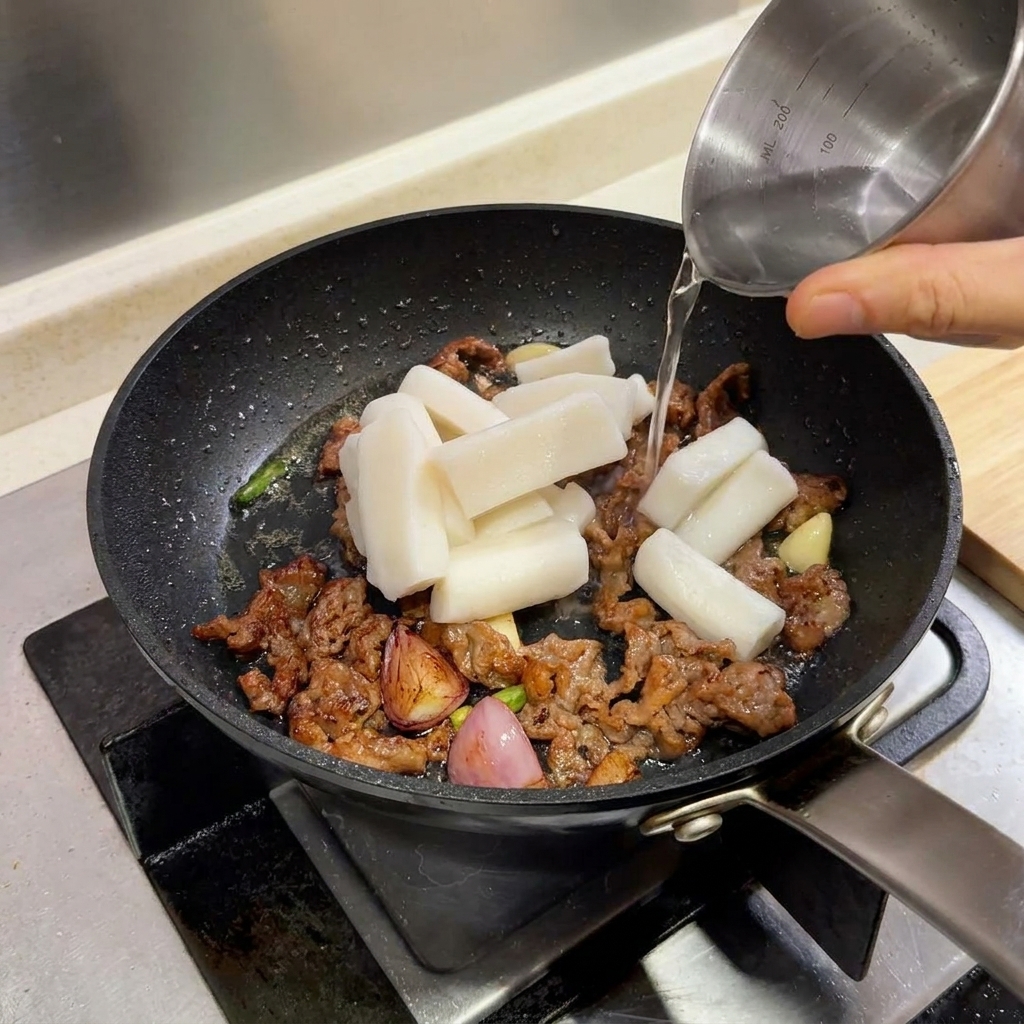 소고기와 함께 프라이팬에 담긴 떡볶이 떡 위로 작은 주전자의 물이 부어지는 모습.