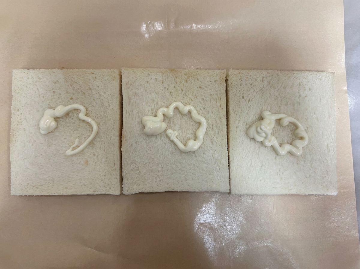 갈색 유산지 위에 나란히 놓인 테두리 없는 흰 식빵 세 장, 각각의 식빵 가운데에 하얀 샐러드 드레싱이 소용돌이 모양으로 짜여 있는 모습.