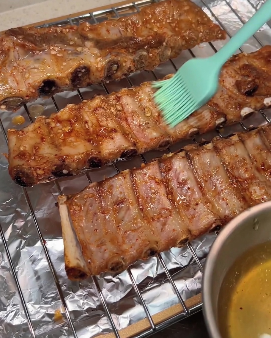 녹색 실리콘 브러시로 식힘망 위의 구워진 돼지갈비 표면에 윤기가 도는 꿀물을 바르는 모습.