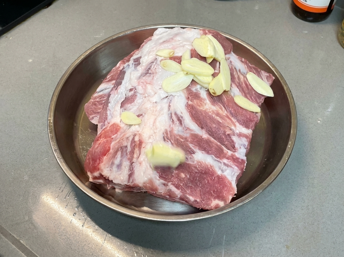 금속 트레이에 놓인 생돼지갈비 위에 마늘 슬라이스가 고르게 흩뿌려진 모습.