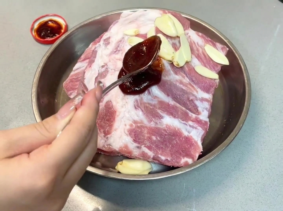 금속 트레이의 돼지갈비 위로 진한 차슈 소스를 숟가락으로 떠서 붓는 모습.