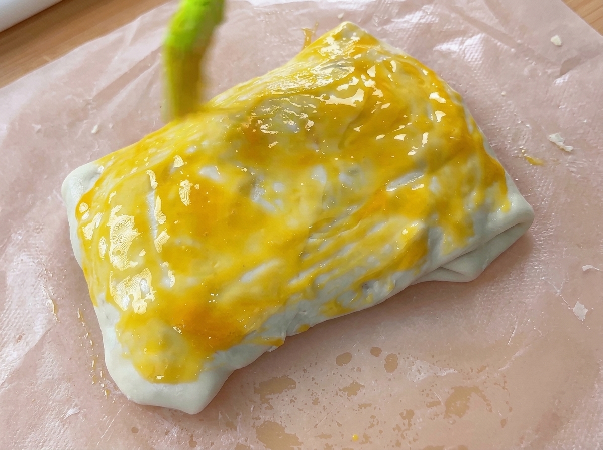 실리콘 브러시로 접힌 반죽 파셀 위에 선명한 노란색 달걀물을 바르는 모습.