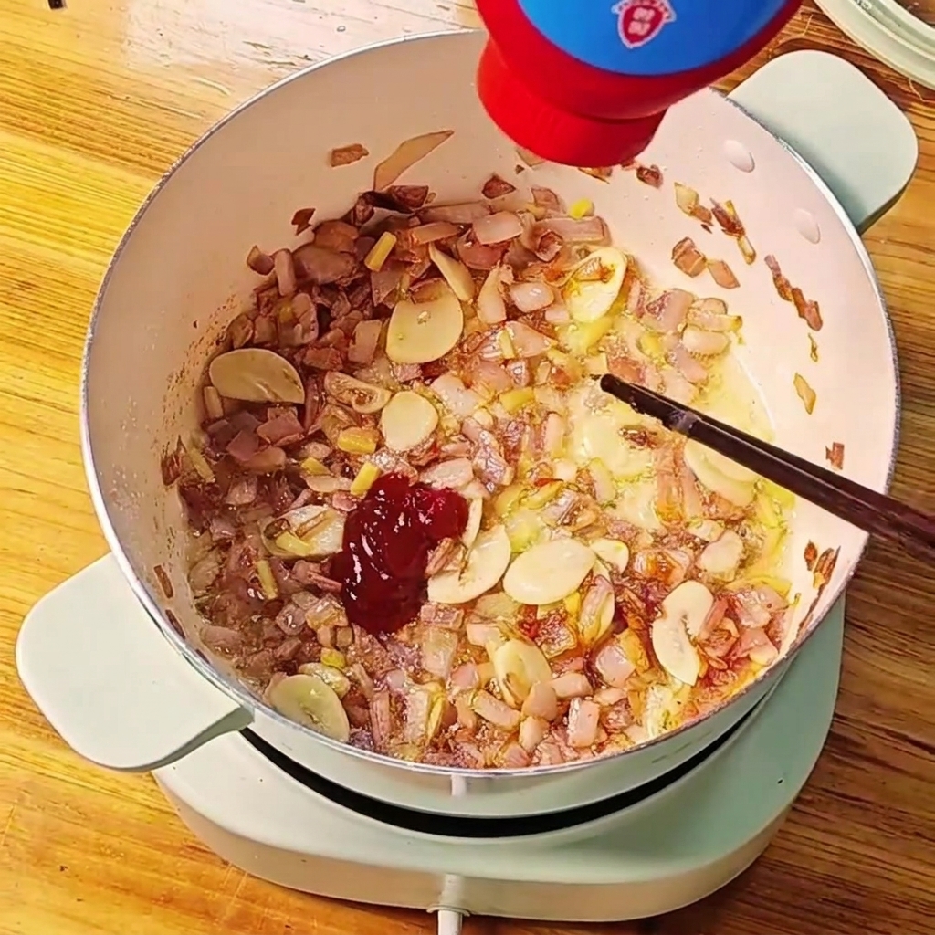 볶은 양파와 마늘이 담긴 냄비에 붉은 토마토 페이스트를 짜 넣는 모습.