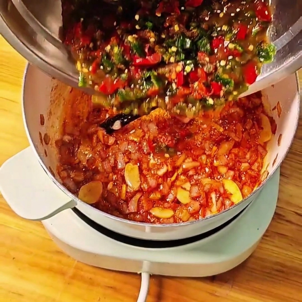 금속 볼에 담긴 찧은 토마토와 고추 혼합물을 조리 중인 냄비에 붓는 모습.