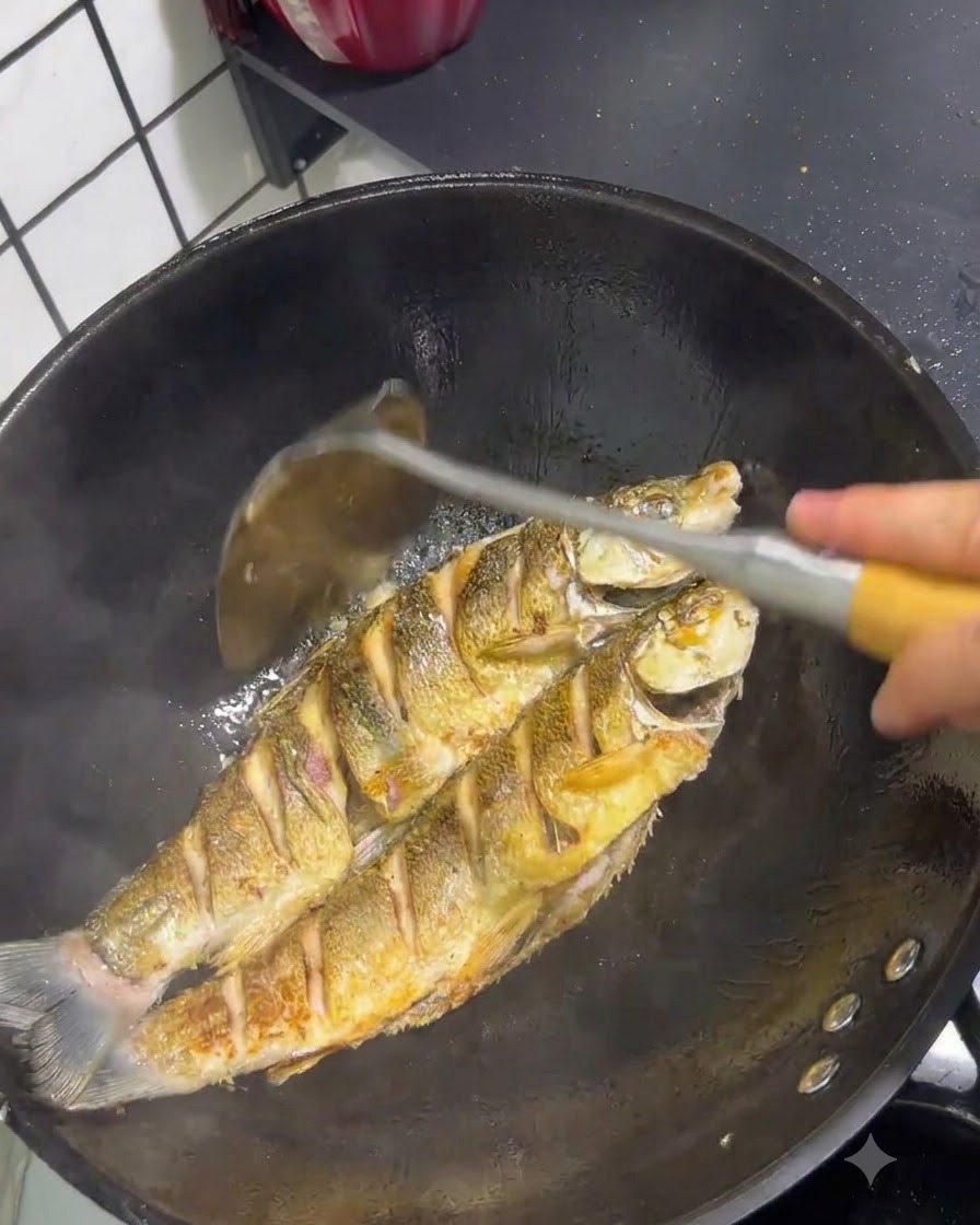 뒤집개로 관리하며 웍 안에서 껍질이 노릇하게 튀겨지고 있는 생선 두 마리.