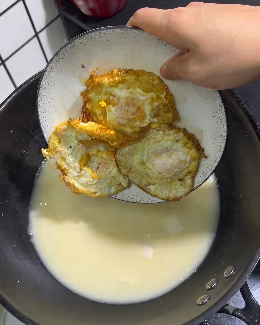 하얀 접시에 담긴 노릇한 달걀프라이 세 개가 보글보글 끓는 국물 속으로 들어가는 모습.