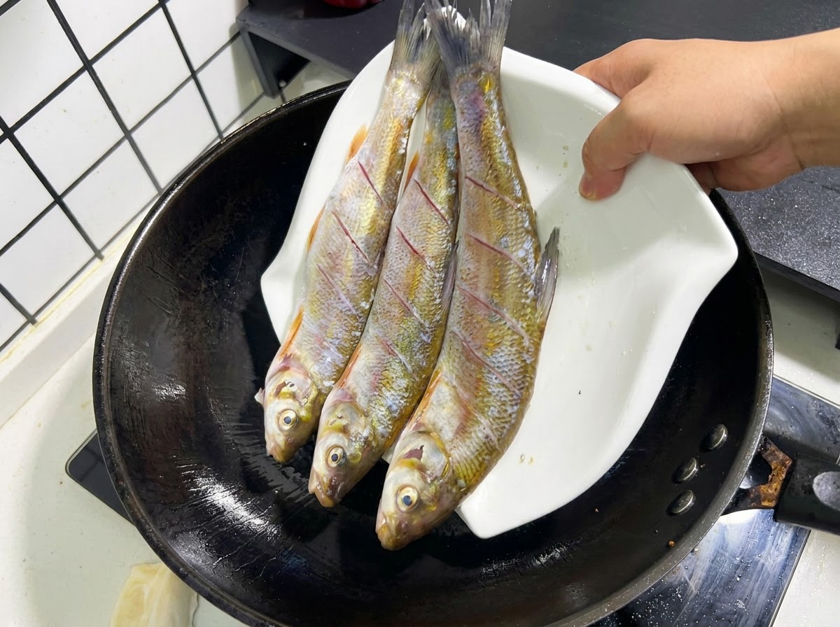 뜨거운 기름이 담긴 검은색 웍에 3마리의 생선을 흰 접시에서 조심스럽게 미끄러지듯 넣는 모습.