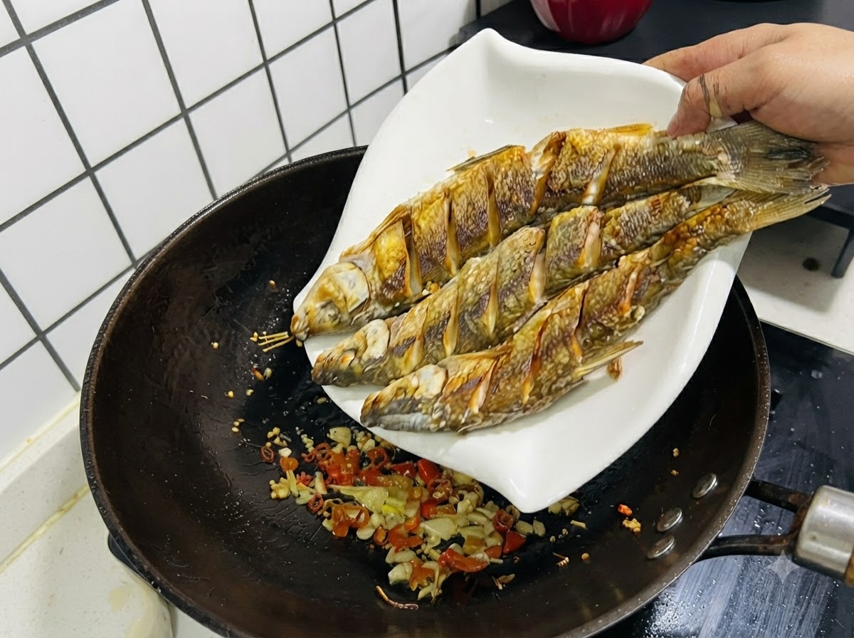 볶은 마늘, 생강, 고추가 들어있는 웍으로 튀긴 생선 세 마리를 흰 접시에서 미끄러지듯 다시 넣는 모습.
