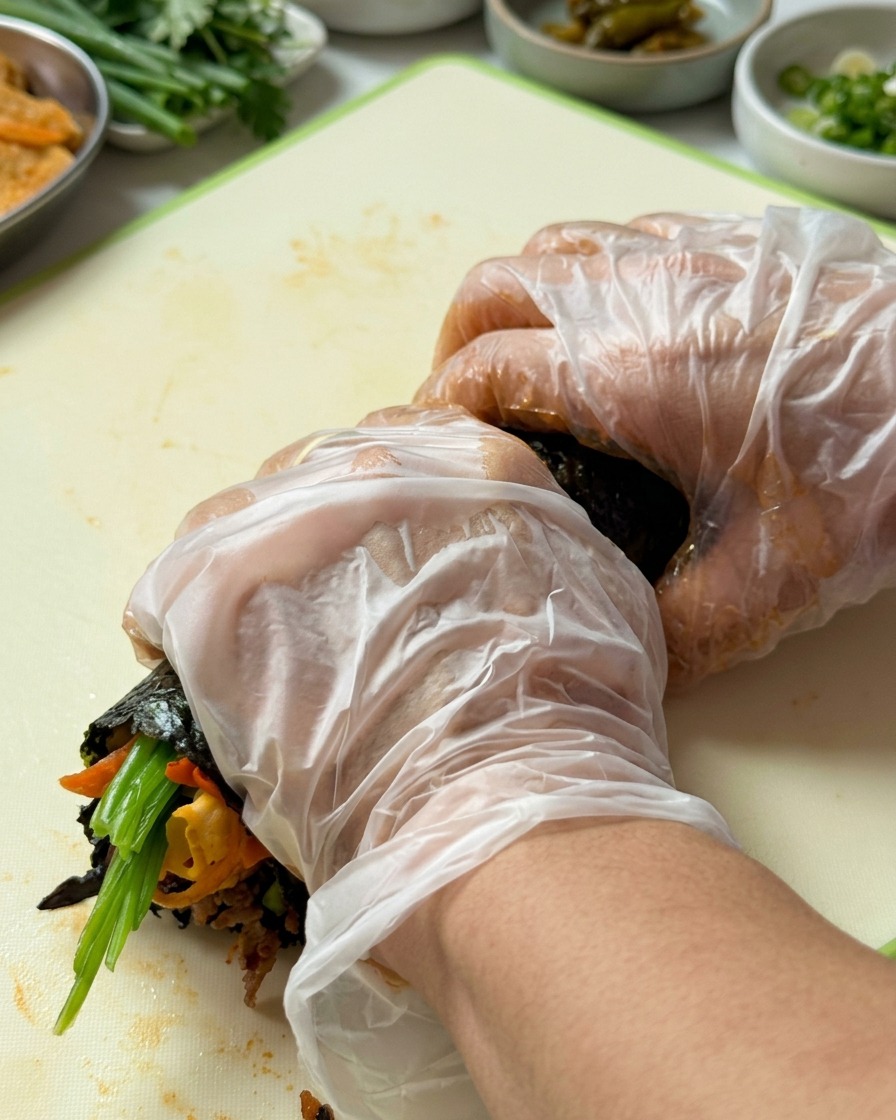 장갑을 낀 손으로 아래쪽부터 위로 김밥을 단단하게 말아 올리는 모습.