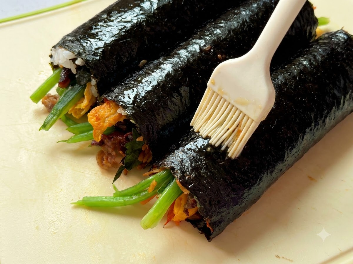 도마 위에 놓인 세 줄의 완성된 김밥에 참기름을 붓칠하는 클로즈업 뷰.