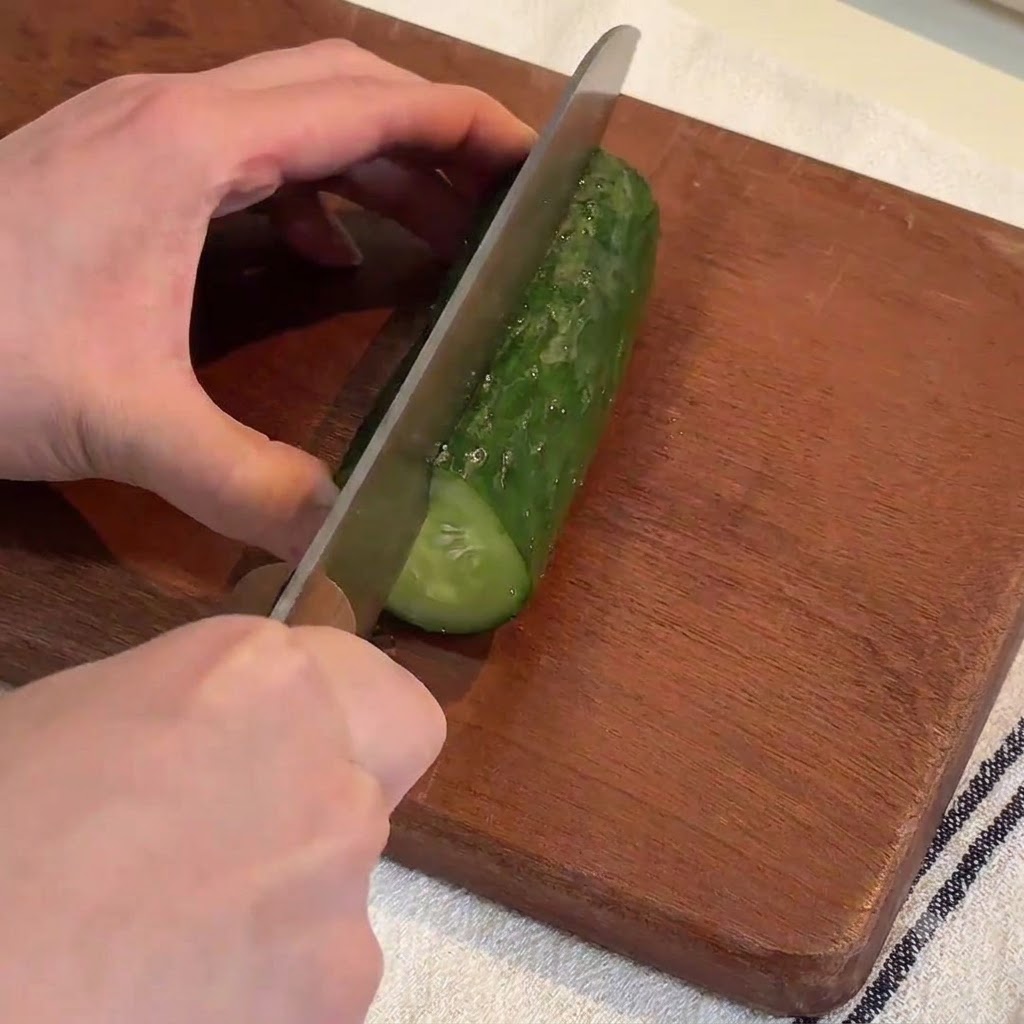 나무 도마 위에서 큰 칼로 신선한 녹색 오이를 긴 조각으로 썰고 있는 손.