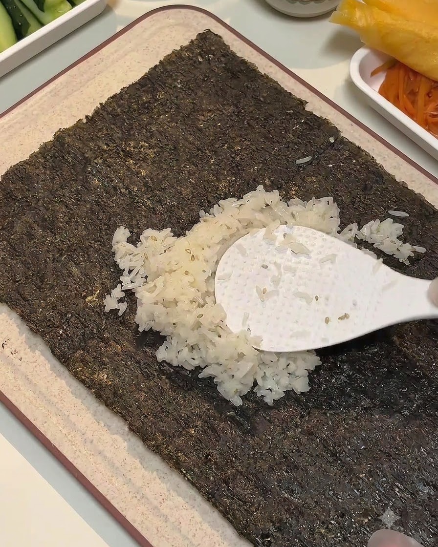 하얀 주걱으로 김 시트 중앙에 양념된 밥을 넓게 펴 바르고 있는 모습.