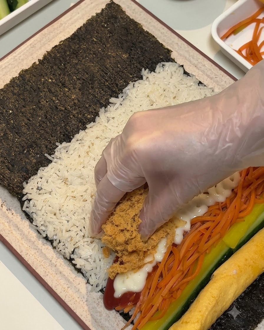 장갑을 낀 손으로 갈색의 폭신한 고기 가루와 햄 스틱을 김 위의 속재료 위로 올리는 모습.