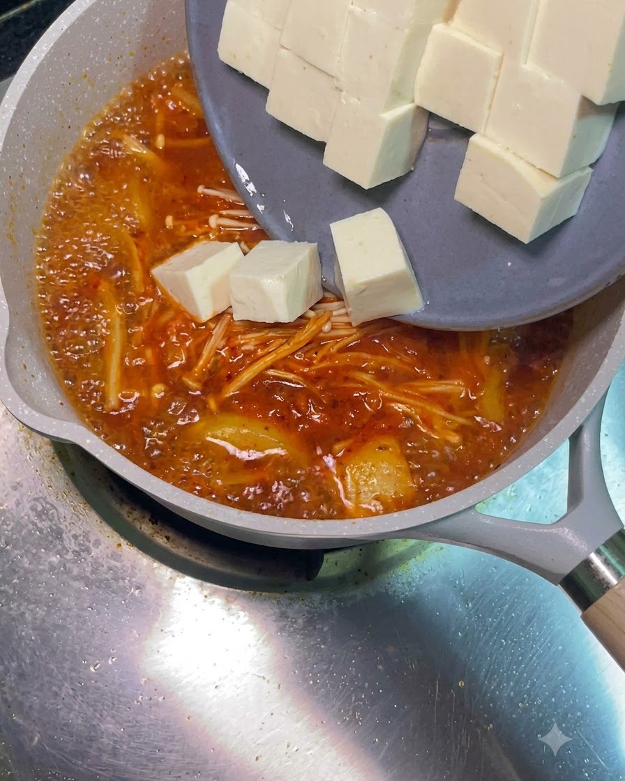 회색 접시에서 네모난 하얀 두부 조각들이 끓고 있는 붉은 찌개 속으로 미끄러져 들어가는 모습.