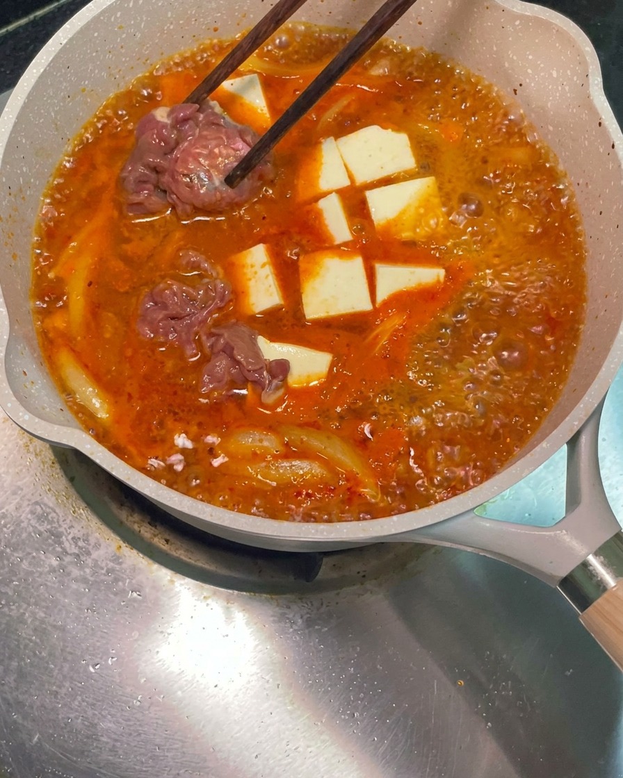 젓가락을 이용해 생고기 슬라이스를 끓고 있는 붉은 찌개 팬에 넣는 모습.