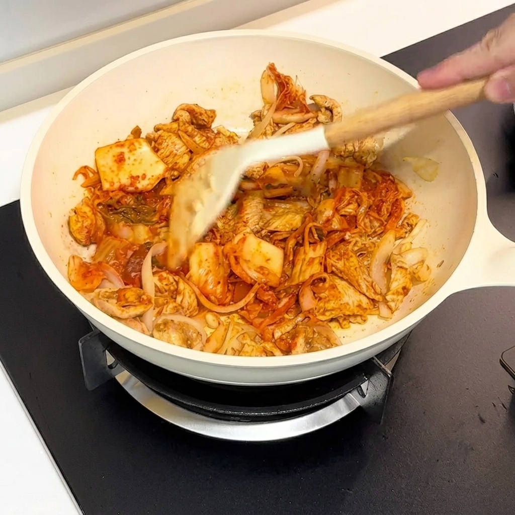 주걱을 사용해 하얀 팬에서 김치와 닭고기를 함께 볶는 모습.