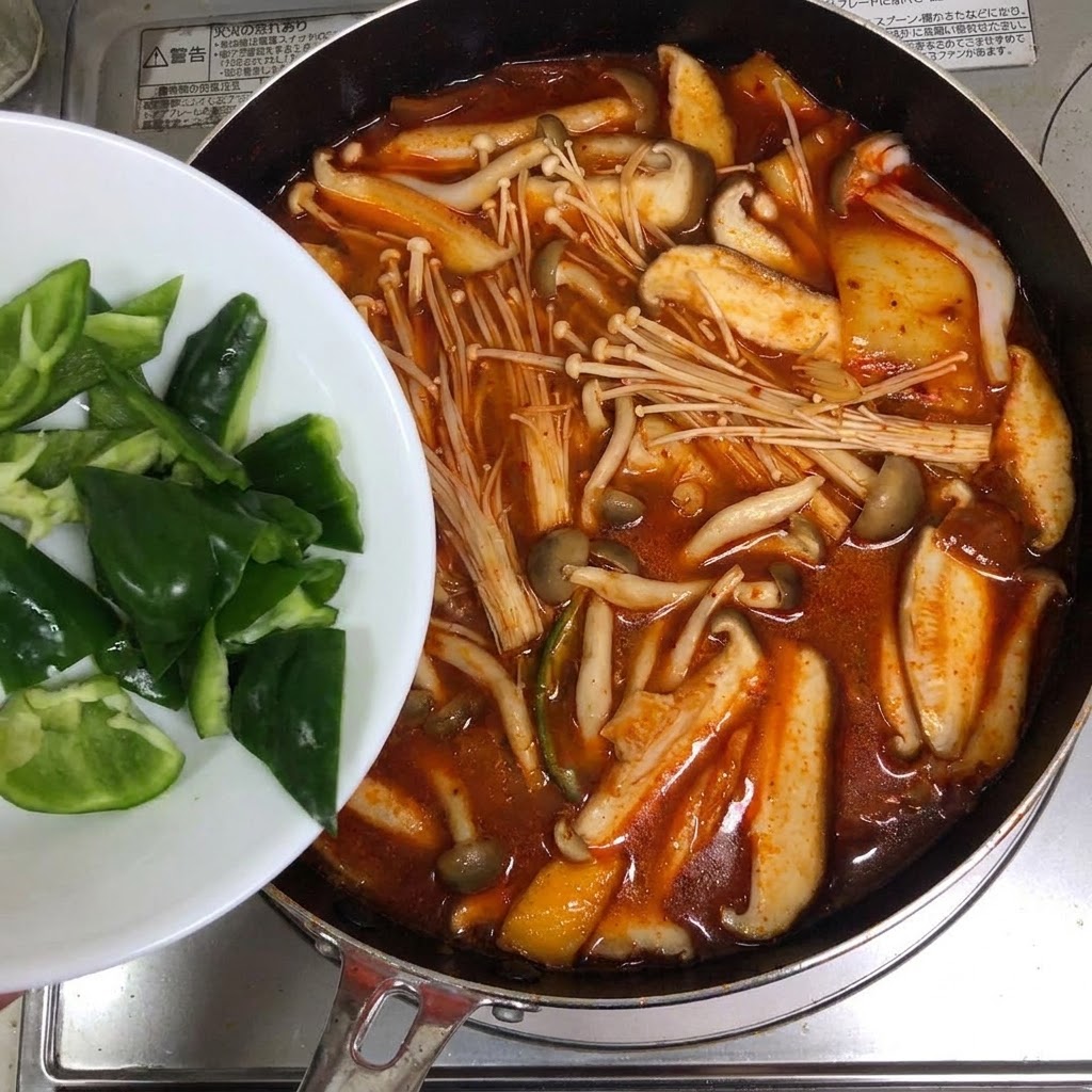 하얀 접시에 담긴 풋고추 슬라이스를 붉은빛의 김치찌개 냄비에 넣는 모습.