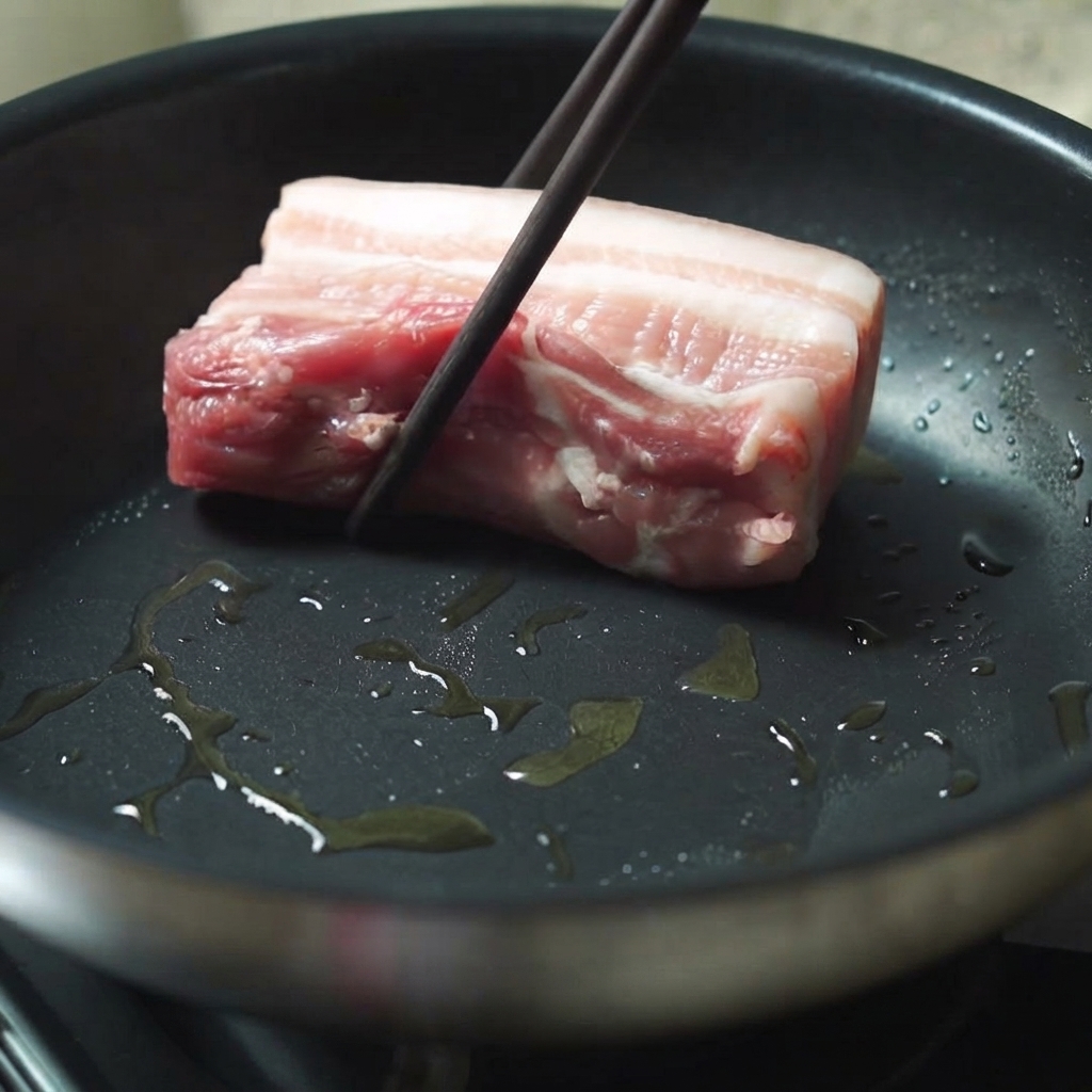 젓가락을 이용해 기름이 달궈진 프라이팬에 두툼한 삼겹살 조각을 넣는 모습.