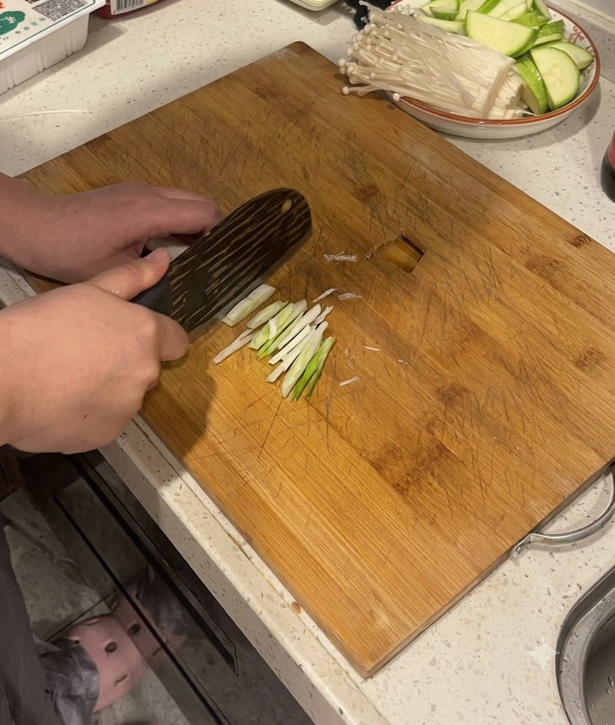 나무 도마 위에서 주방용 칼을 사용하여 대파 줄기를 썰고 있는 손.