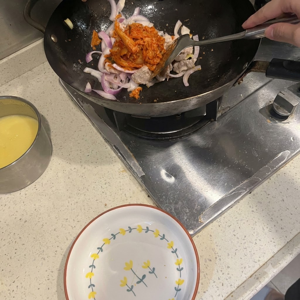 볶아진 삼겹살과 적양파가 담긴 웍에 주걱으로 새빨간 김치를 넣는 모습.