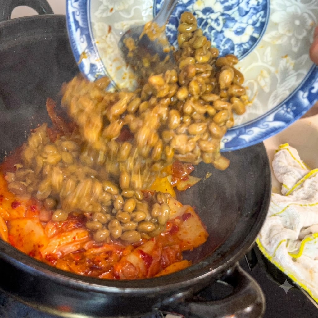 파란색과 흰색이 섞인 무늬의 접시에서 발효된 콩이 지글지글 끓는 김치 냄비 속으로 미끄러져 들어가는 모습.
