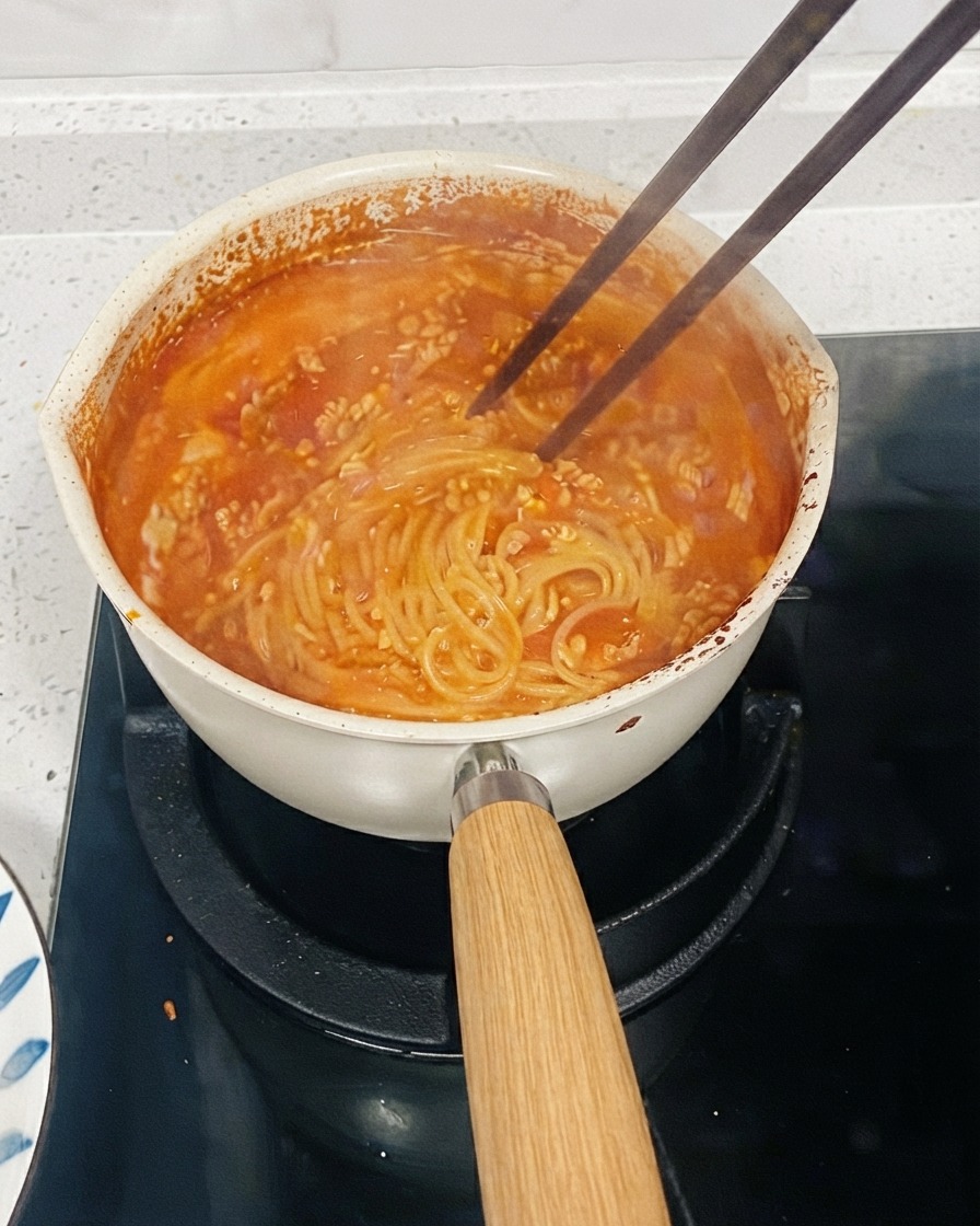 소스를 걸쭉하게 만들기 위해 젓가락으로 면을 저어가며 스토브 위에서 끓고 있는 토마토 파스타 냄비.
