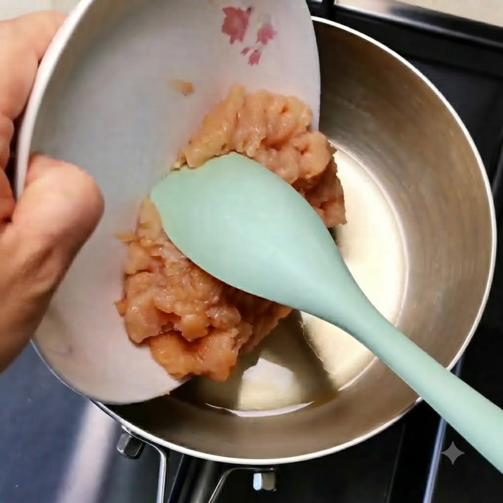 초록색 주걱을 이용해 하얀 접시에 담긴 다진 닭고기를 예열된 기름 팬으로 밀어 넣는 모습.
