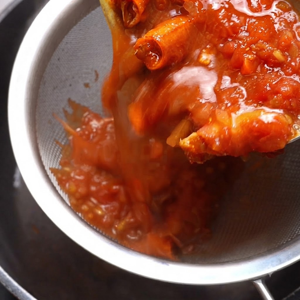 그릇 위에 올린 금속 체에 익은 토마토 소스를 붓고 주걱으로 건더기를 누르는 모습.