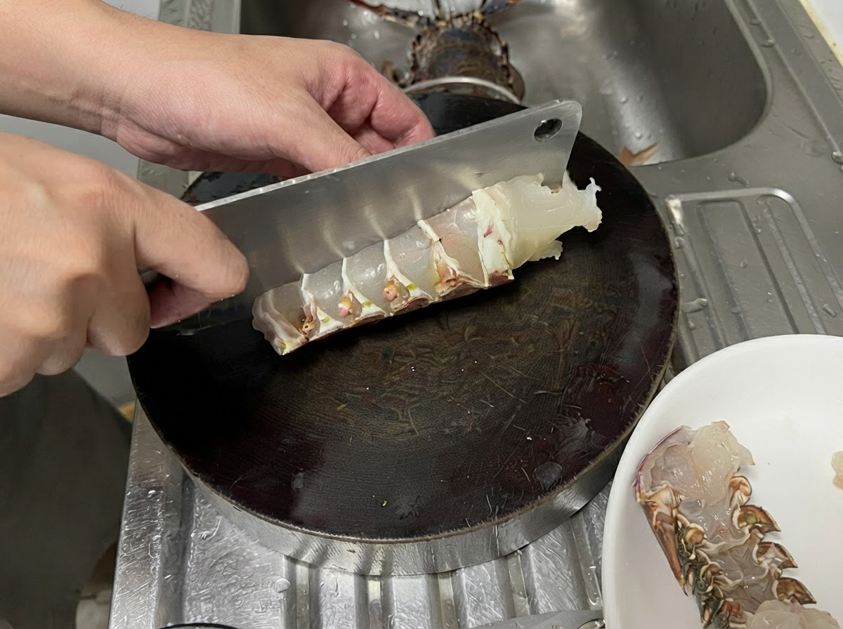어두운 원형 도마 위에서 큰 중식도로 날것의 랍스터 꼬리를 썰어 두껍고 고른 조각을 만드는 모습.