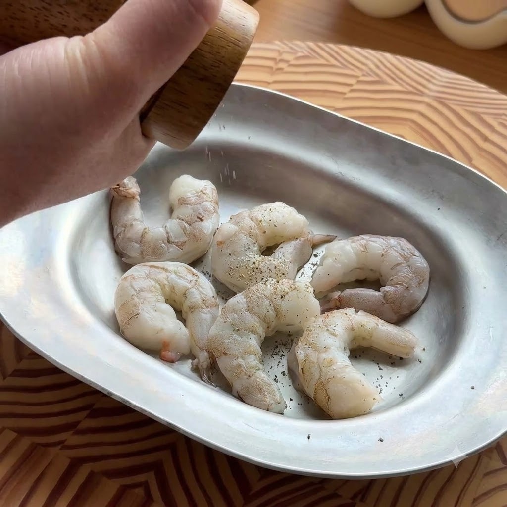 타원형 메탈 트레이에 담긴 손질된 생새우에 나무 페퍼 그라인더로 후추를 갈아 넣는 손.