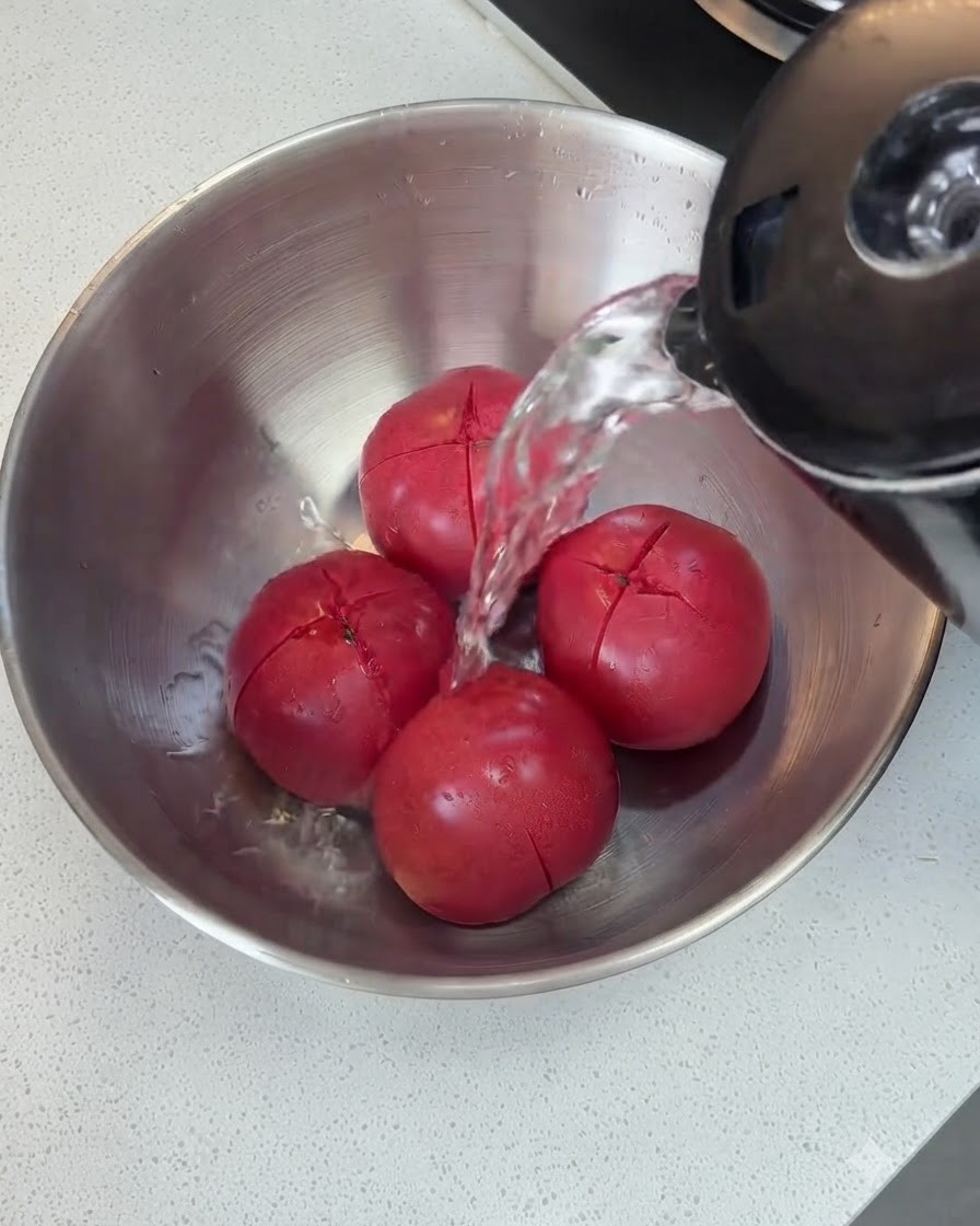 주전자에서 끓는 물이 금속 볼 안의 칼집 낸 토마토 위로 뜨겁게 부어지는 모습.