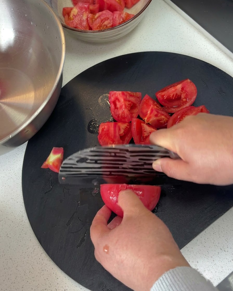 어두운 도마 위에서 칼을 이용해 껍질 벗긴 토마토를 먹기 좋은 크기의 덩어리로 썰고 있는 모습.
