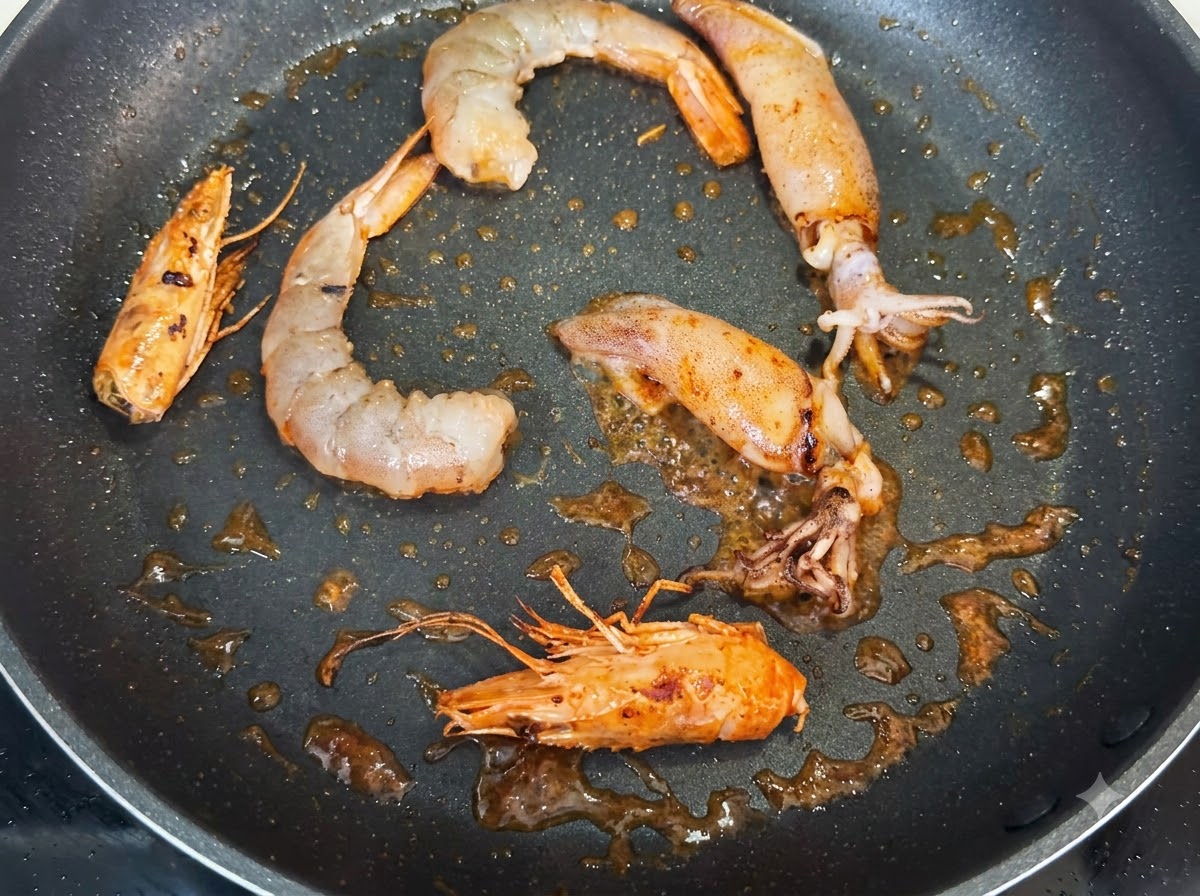 팬에서 껍질 깐 새우, 통새우 머리, 오징어 조각들이 검은 후추 가루와 함께 볶아지는 모습.