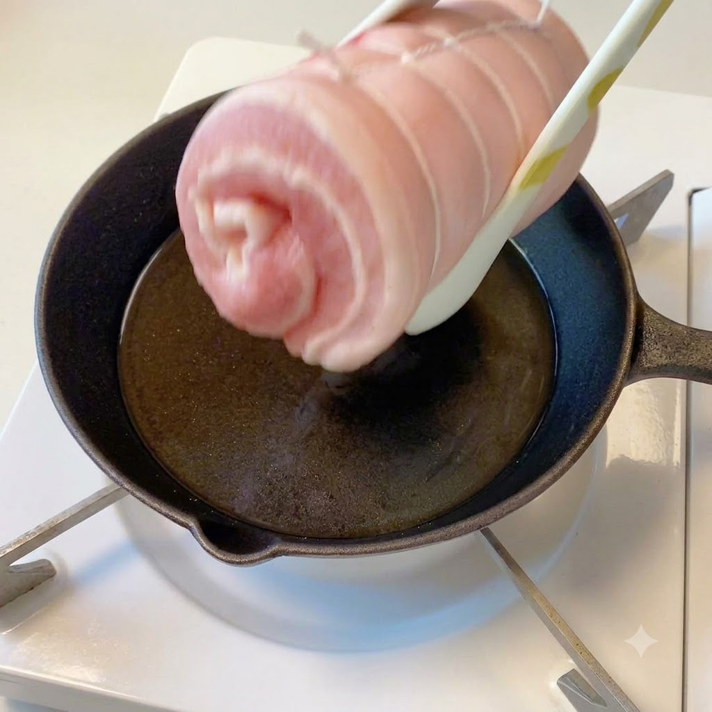 집게로 집은 실로 묶인 삼겹살 실린더를 기름이 달궈진 프라이팬 위에서 익히는 모습.