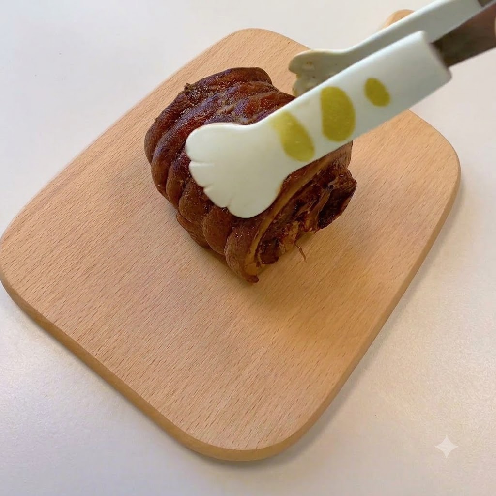 나무 도마 위에 올려진 조리된 롤 형태의 차슈를 집게로 잡고 있는 모습.