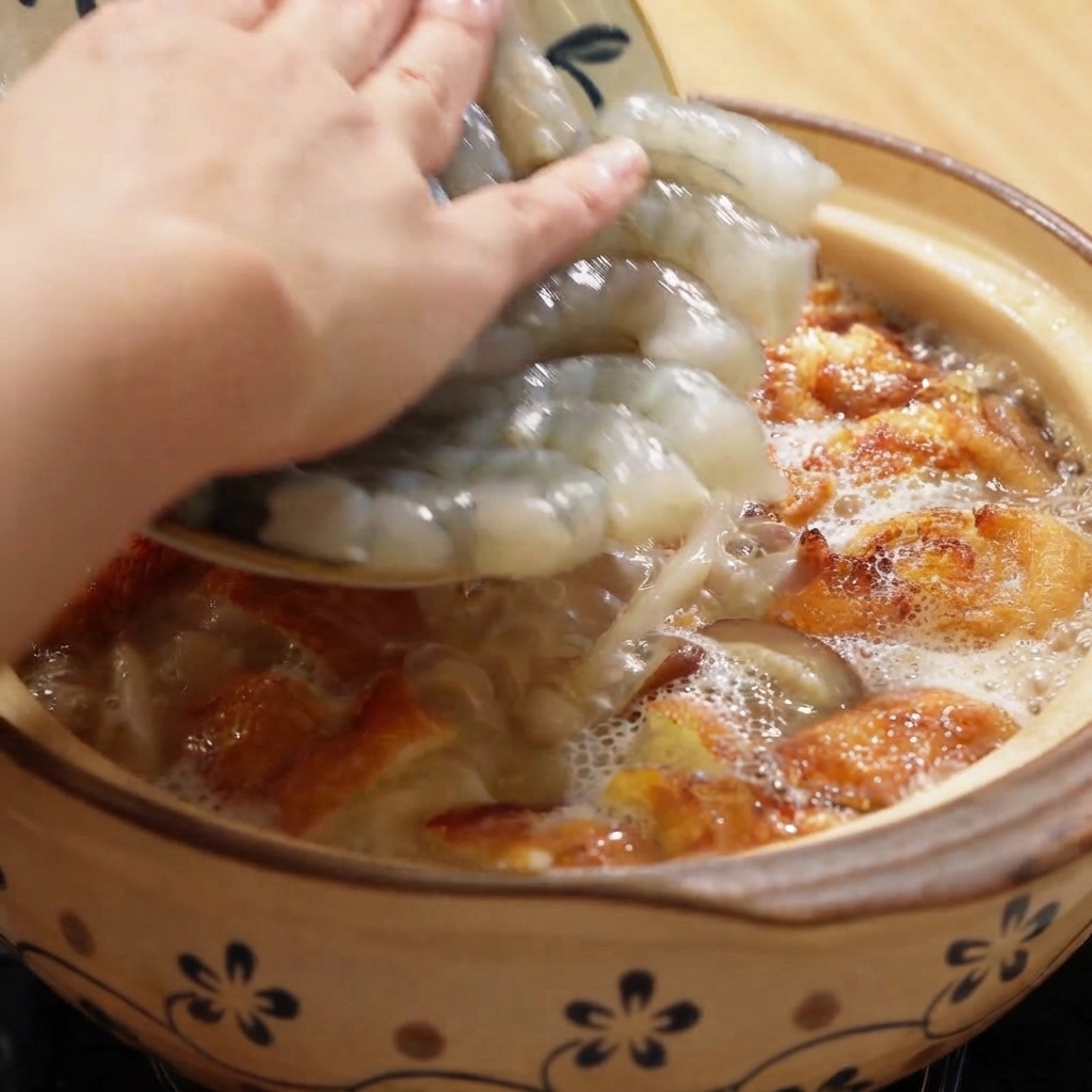 국물과 달걀 조각이 가득 찬 보글보글 끓는 뚝배기에 껍질 깐 새우를 넣는 모습.