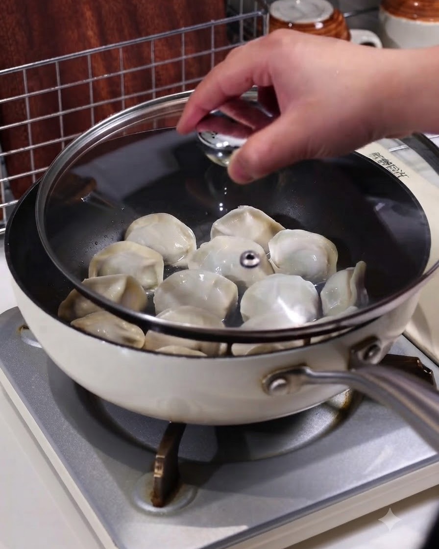 스토브 위에서 만두가 찌어지고 있는 팬에 투명한 유리 뚜껑을 덮고 있는 손의 모습.