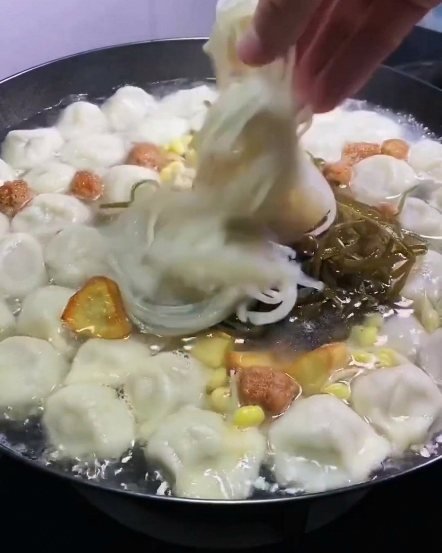 끓는 만두와 육수가 담긴 냄비에 채 썬 녹색 미역과 투명한 당면을 손으로 넣는 모습.