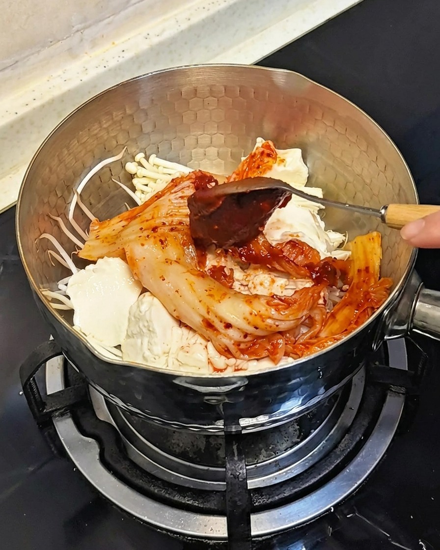 냄비 안의 재료 위에 숟가락으로 붉은 고추장을 얹는 모습.
