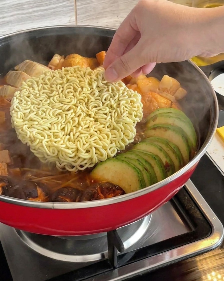 끓고 있는 한국식 부대찌개 중앙에 손으로 건조 인스턴트 라면 면사리를 넣는 모습.